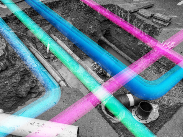 Image of a hole in the ground showing pipes and cables with a pink, blue and green coloured lines that look like pipes sitting on the surface..